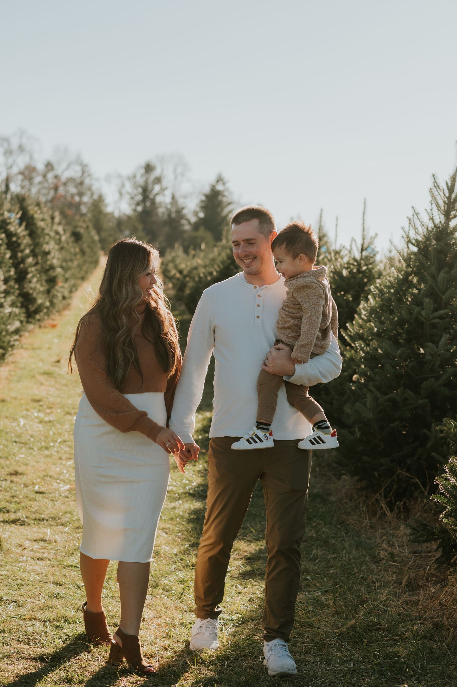Young family with baby walks through sunny Christmas tree farm in casual coordinated outfits.