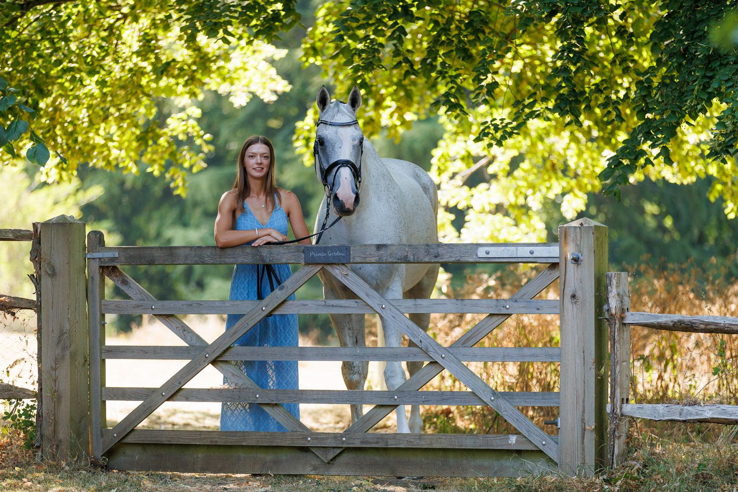 Young woman in black top smiling while gently embracing a brown horse outdoors among lush green trees.