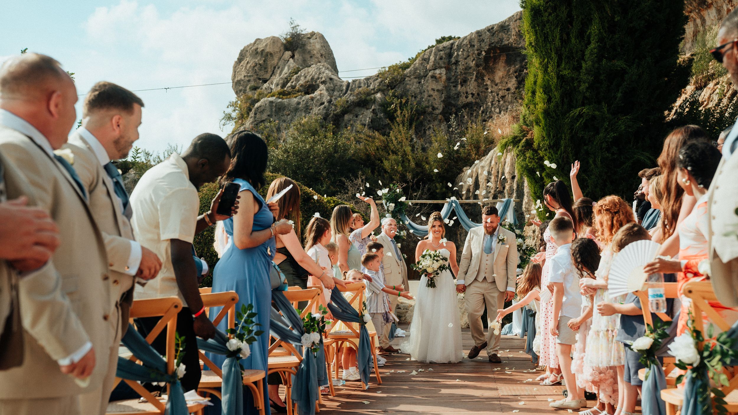Bride in a white gown walks down an outdoor aisle between rows of seated wedding guests with a rocky cliff and greenery in the background.