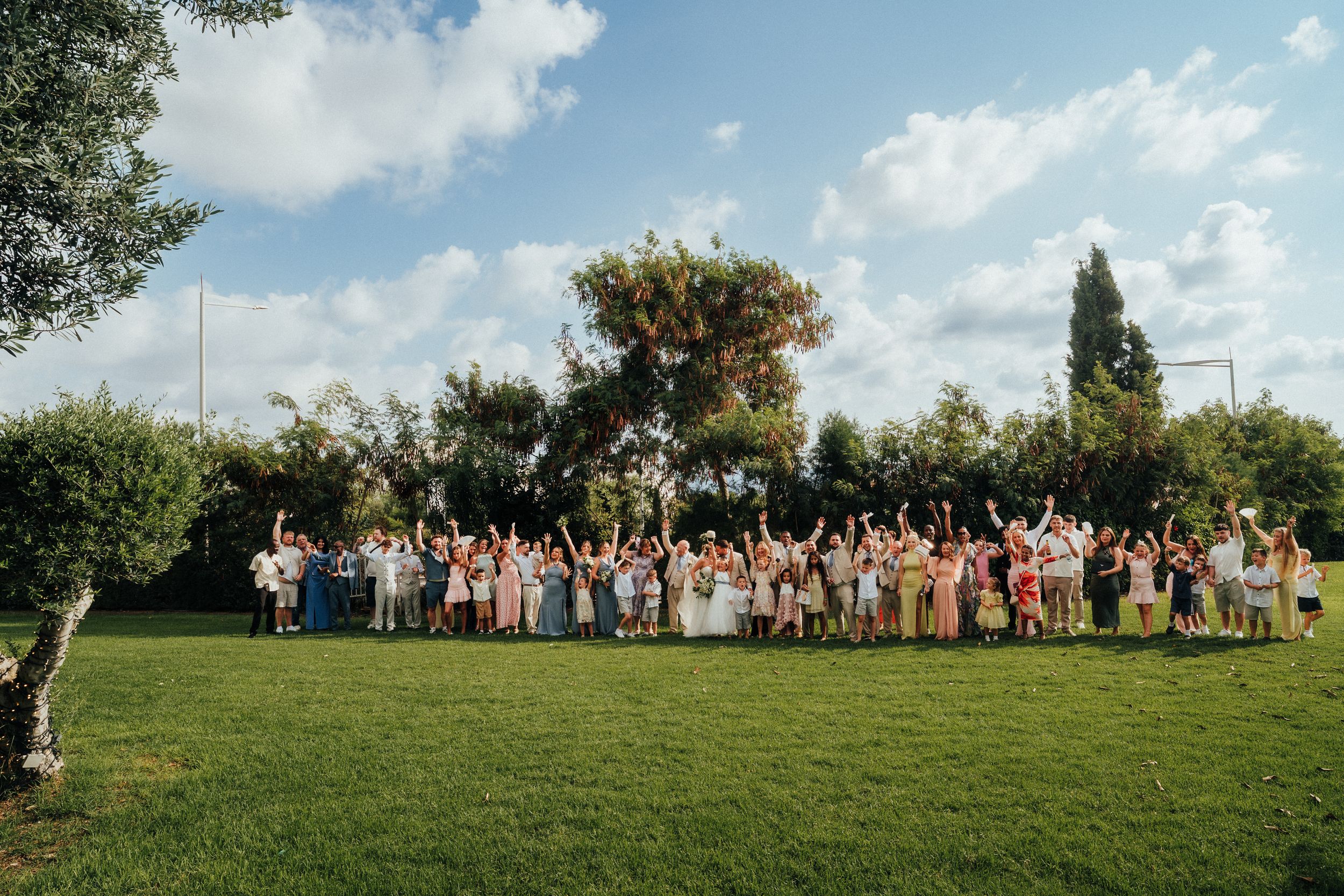 Large group of wedding guests in colorful attire cheering with raised arms on a sunny green lawn surrounded by trees under a blue sky.