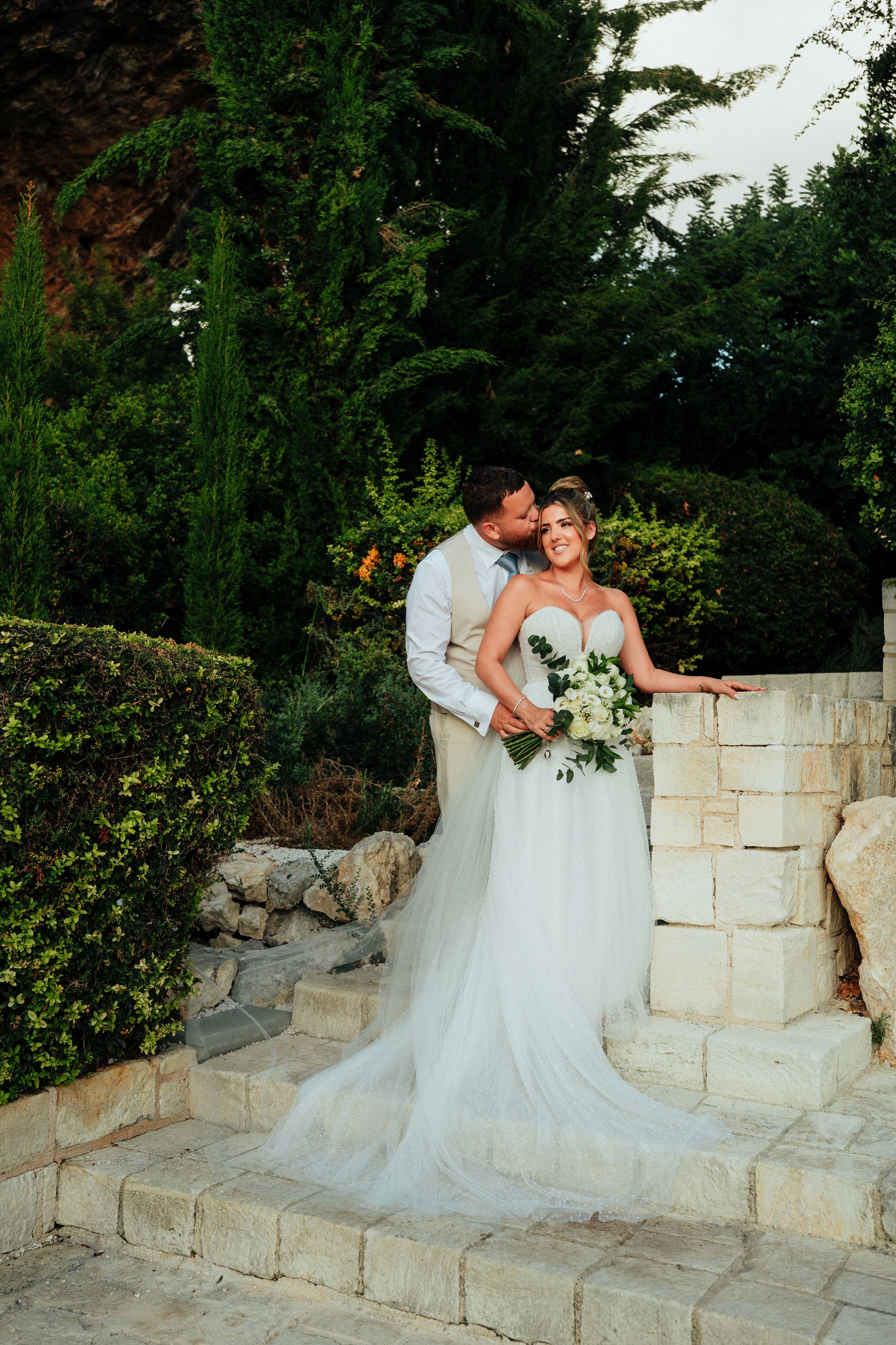 Groom in a vest kisses bride in a tulle wedding gown holding a bouquet on stone steps surrounded by greenery.