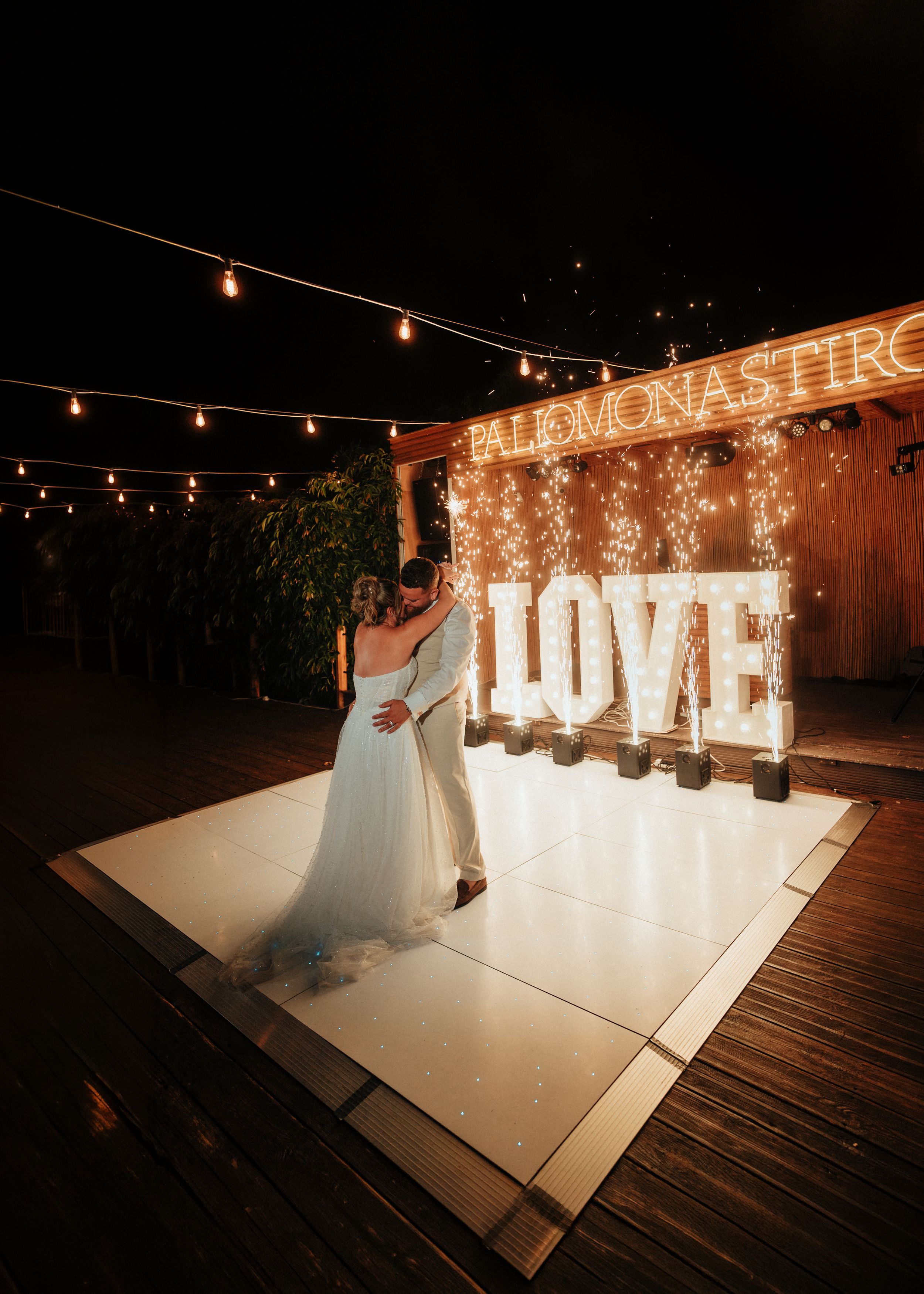 Bride and groom share a romantic first dance on a white dance floor beside glowing marquee LOVE letters under twinkling string lights.