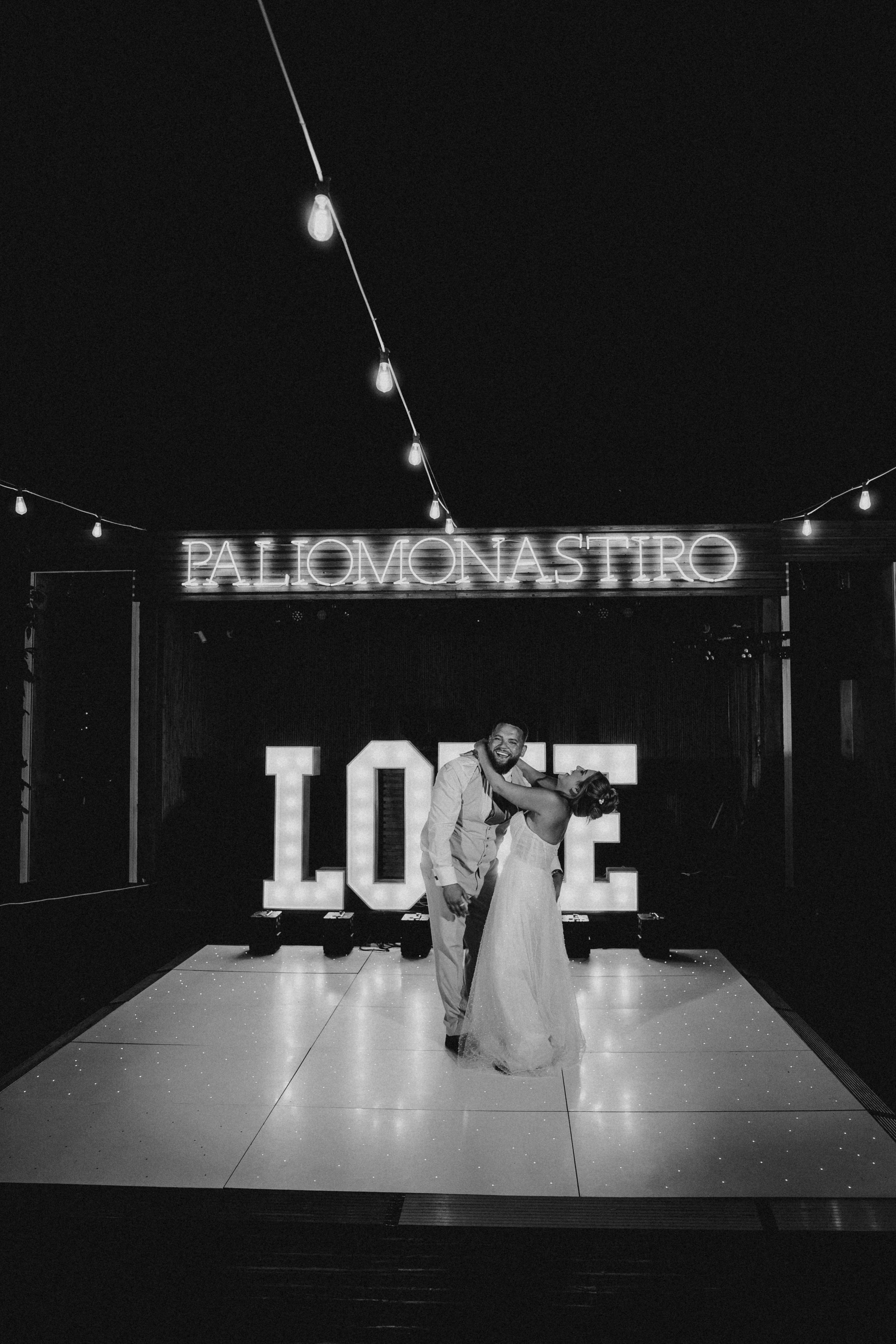 Black and white photo of a couple sharing a dance on a lit dance floor in front of a glowing LOVE sign at a Paliomonastiro wedding.