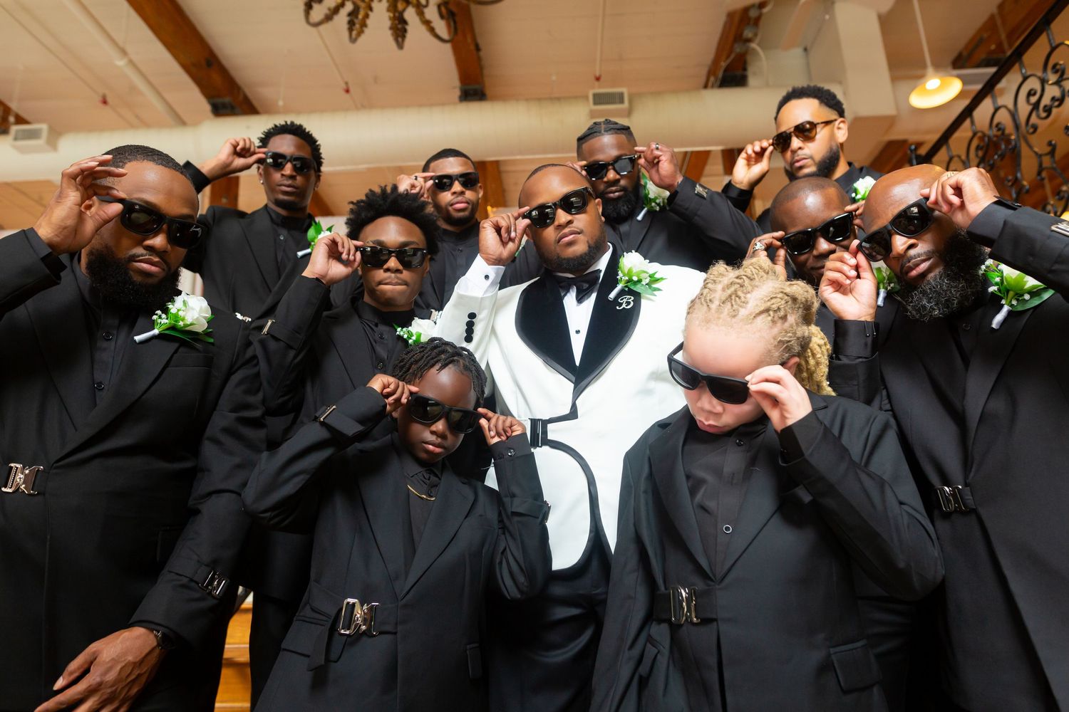 Groomsmen in black suits and sunglasses strike a playful pose around a groom in a white tuxedo jacket at a wedding venue.