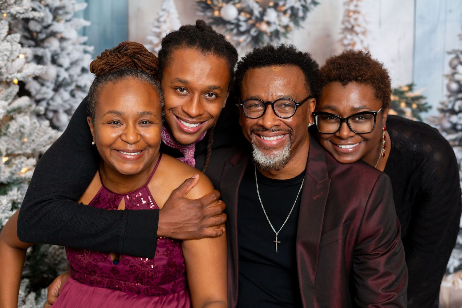 Four smiling people in burgundy and black outfits pose close together in a joyful family portrait in front of frosted holiday trees.