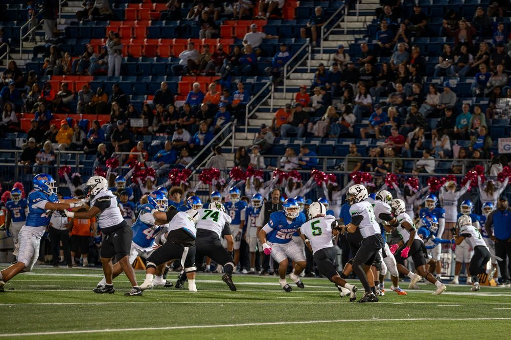 Football players in white and blue uniforms clash on a green field while fans fill the stadium bleachers during a night game.