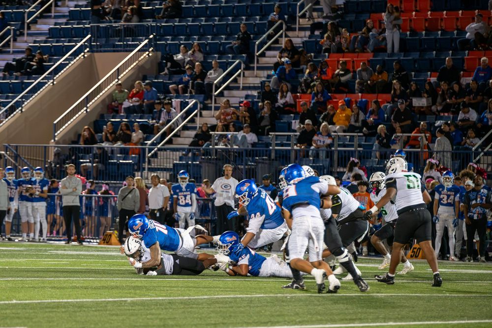 Football players in blue and white uniforms tackle a ball carrier on the field during a night game as fans watch from the stadium stands.