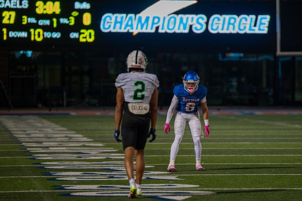 Two high school football players, one in a white number 2 jersey and one in blue, face off on a turf field under stadium lights.