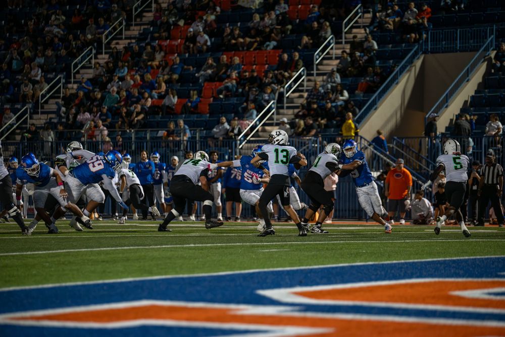 Football players in blue and white uniforms clash at the line of scrimmage during a night game as fans watch from the stadium stands.