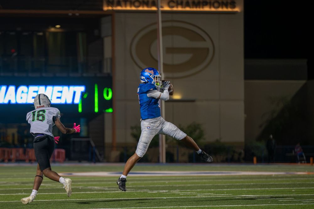 A football player in a blue uniform leaps to catch a pass at night while a defender in a white jersey runs nearby on a stadium field.