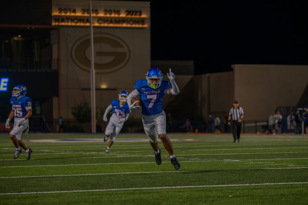 Football player in blue jersey number 7 sprints across the field at night as teammates run behind him near a stadium building.