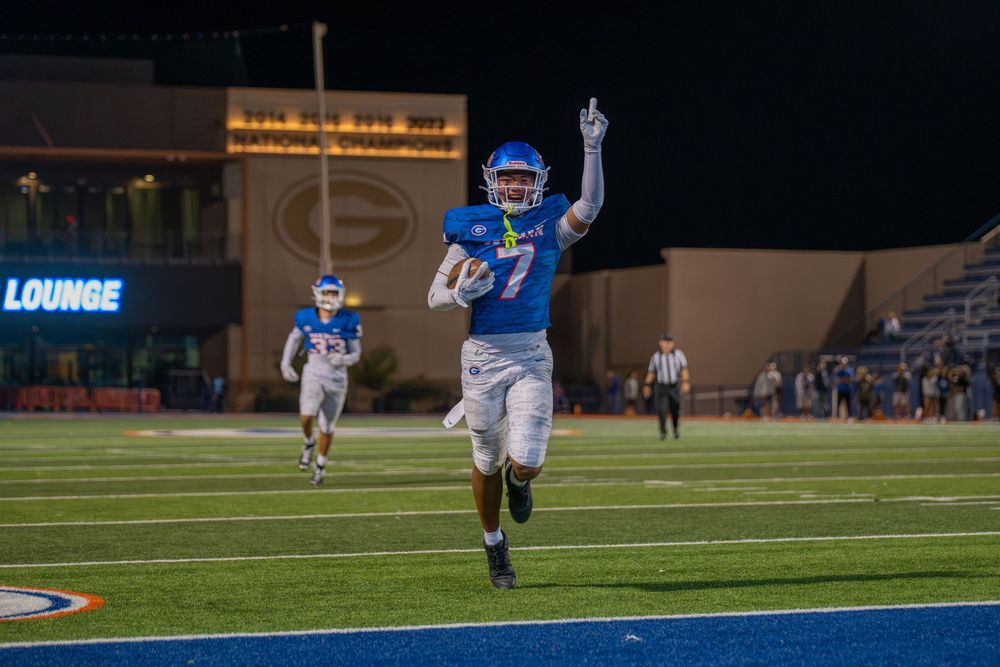 A football player in a blue uniform raises one arm while running down the field at night as a teammate follows behind.