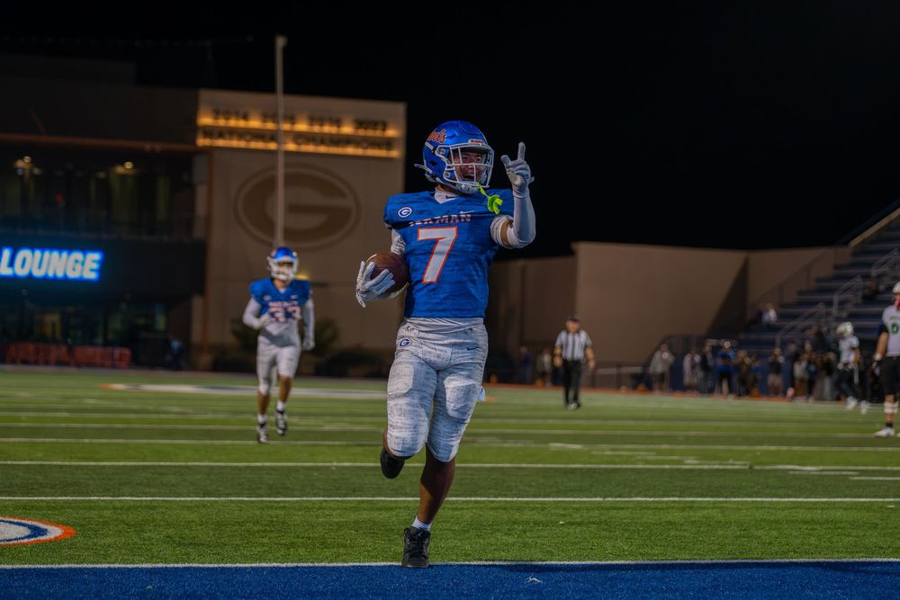 A football player in a blue number 7 jersey and helmet points upward while running with the ball under stadium lights at night.