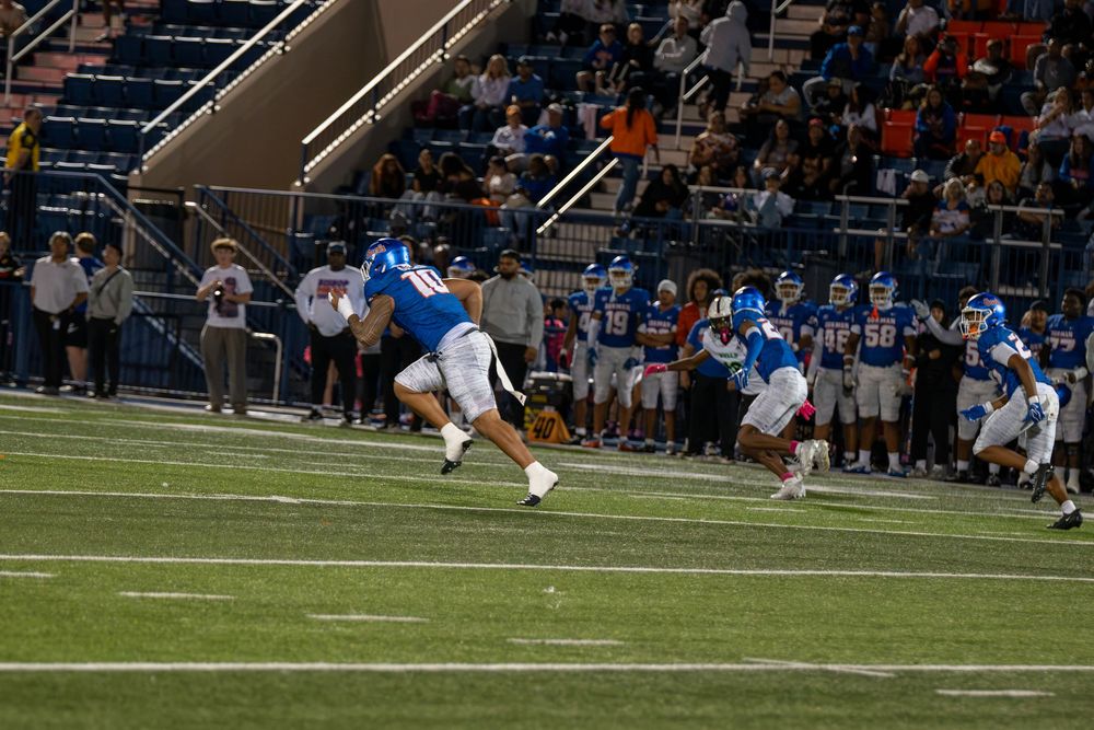 Football player in a blue uniform runs downfield during a night game as teammates and a crowd watch from the sidelines and stands.