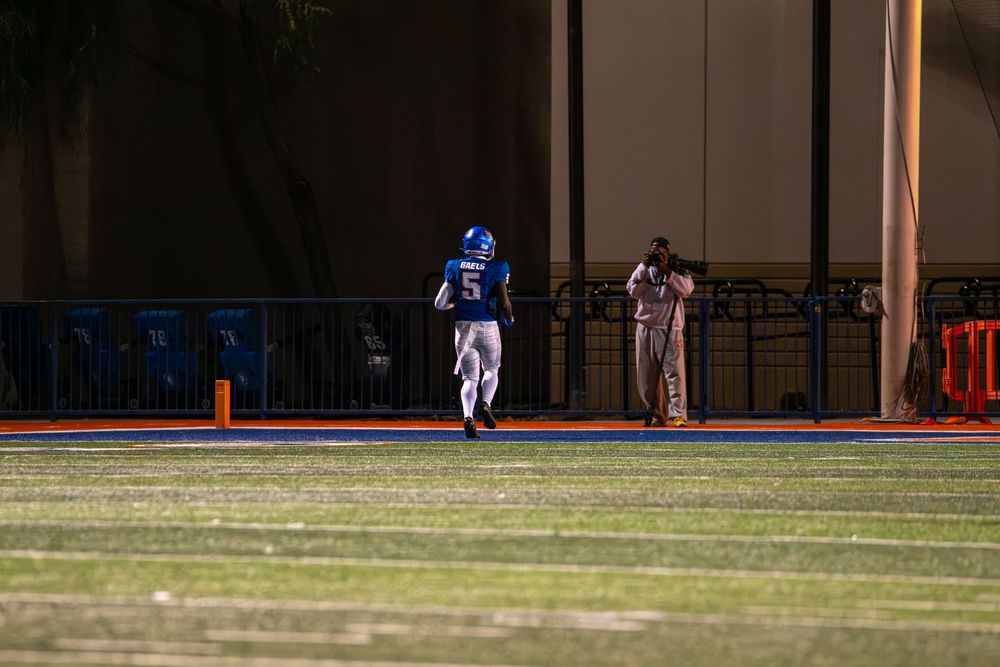 A football player in a blue uniform and helmet runs across the turf field at night while a camera operator films from the sideline.