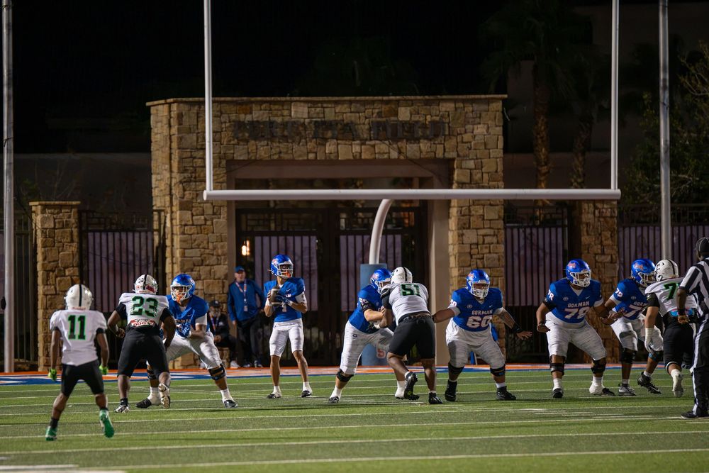Football players in blue and white uniforms line up at the line of scrimmage during a night high school game under stadium lights.