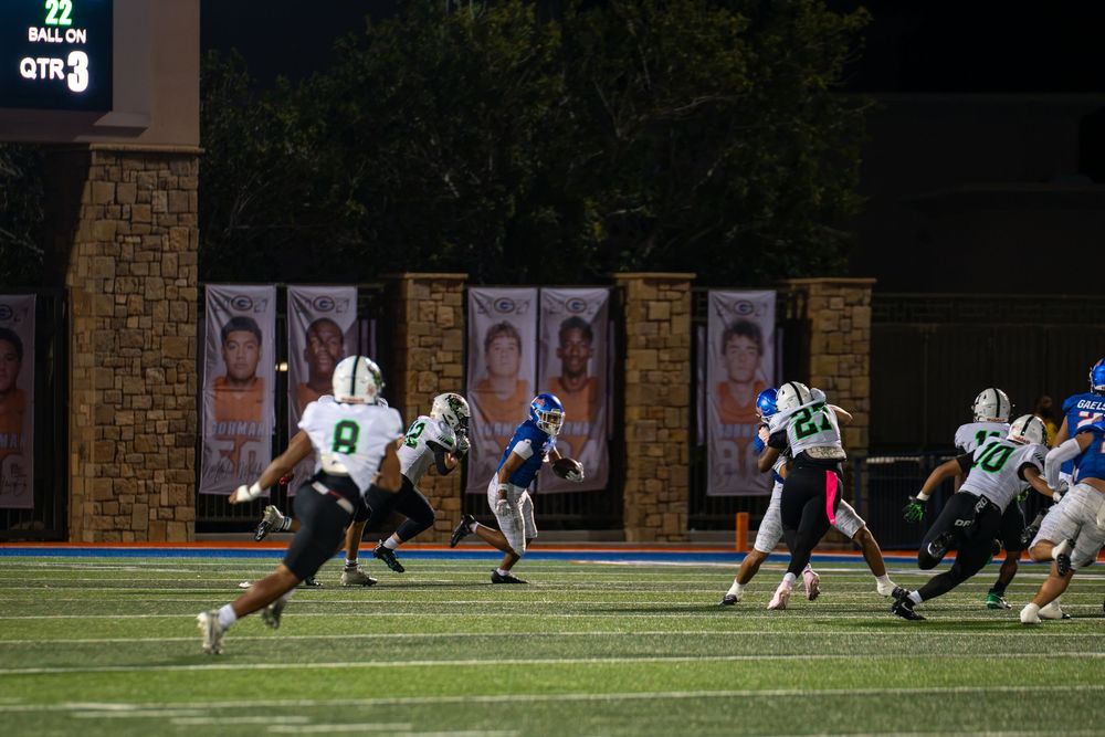 High school football players in white and blue uniforms run across the field during a night game with banners and a scoreboard in the backgr