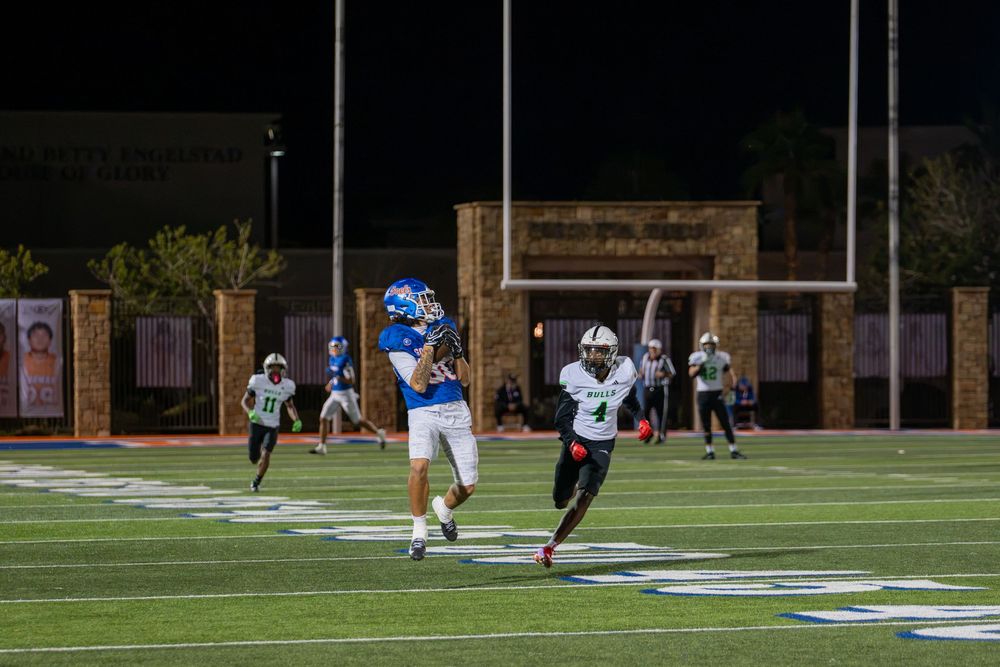 A football player in a blue uniform runs downfield during a night game while teammates and opponents follow near the end zone.