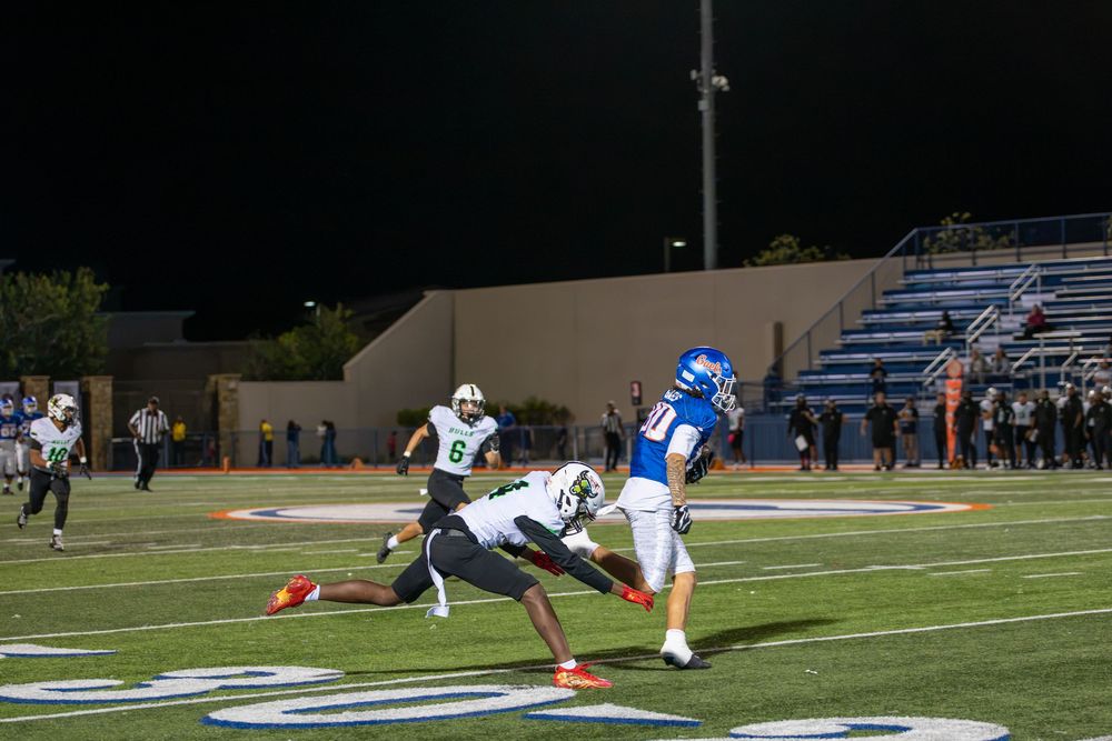 A player in a blue uniform runs downfield as a defender in a dark jersey dives to tackle him during a night high school football game.