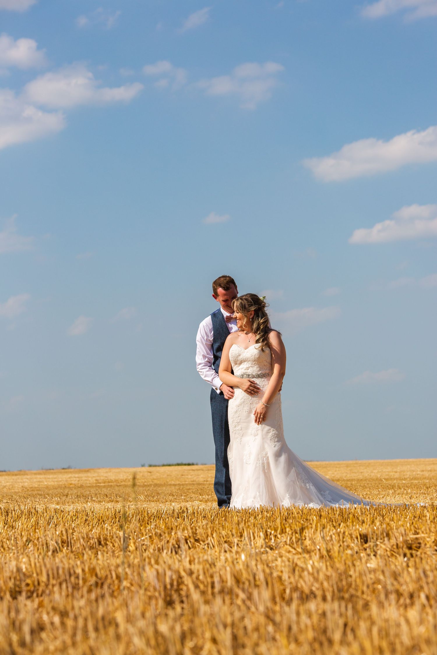 Bride and groom embracing in golden wheat field under blue sky on their wedding day.
