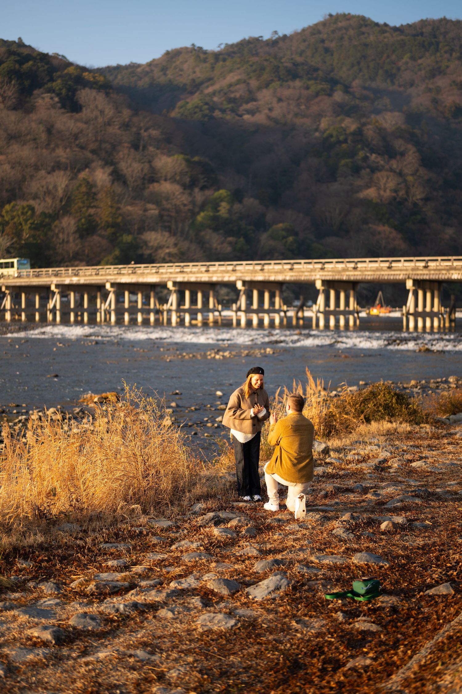 Couple holding hands facing each other on a riverside path beneath pine trees, with a long bridge and forested hills in the background.