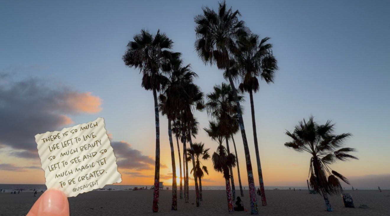 Hand holding a handwritten note in front of silhouetted palm trees on a beach at sunset with a glowing orange and blue sky.