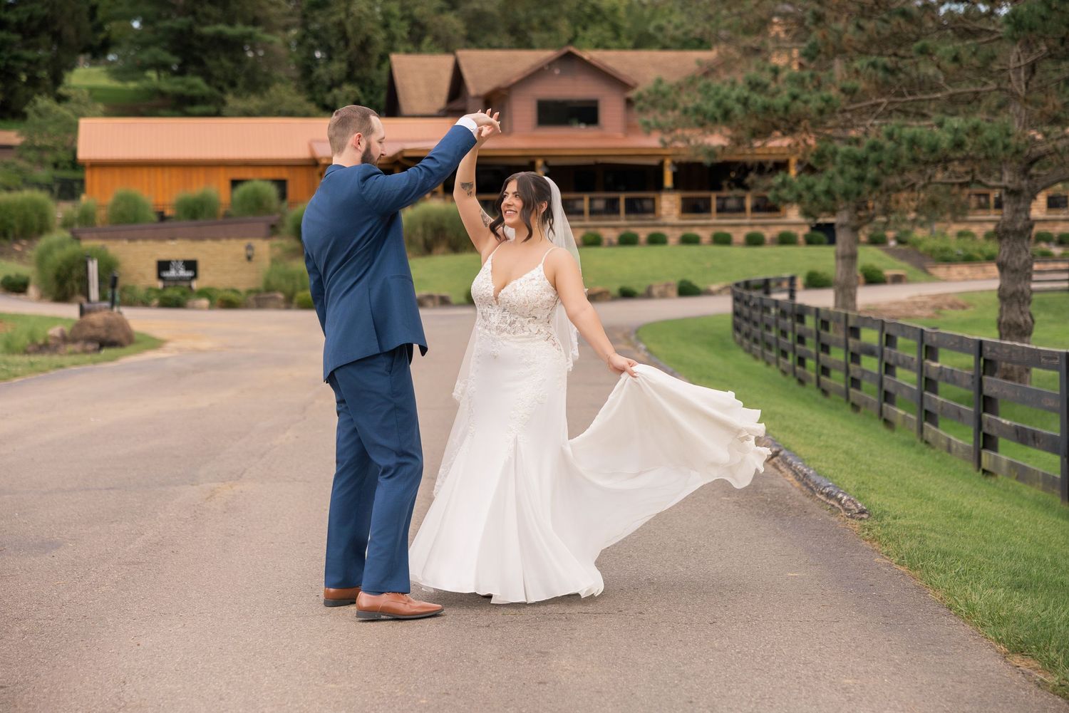 Bride and groom dancing on a driveway outside a rustic wedding venue, bride twirling in white gown.