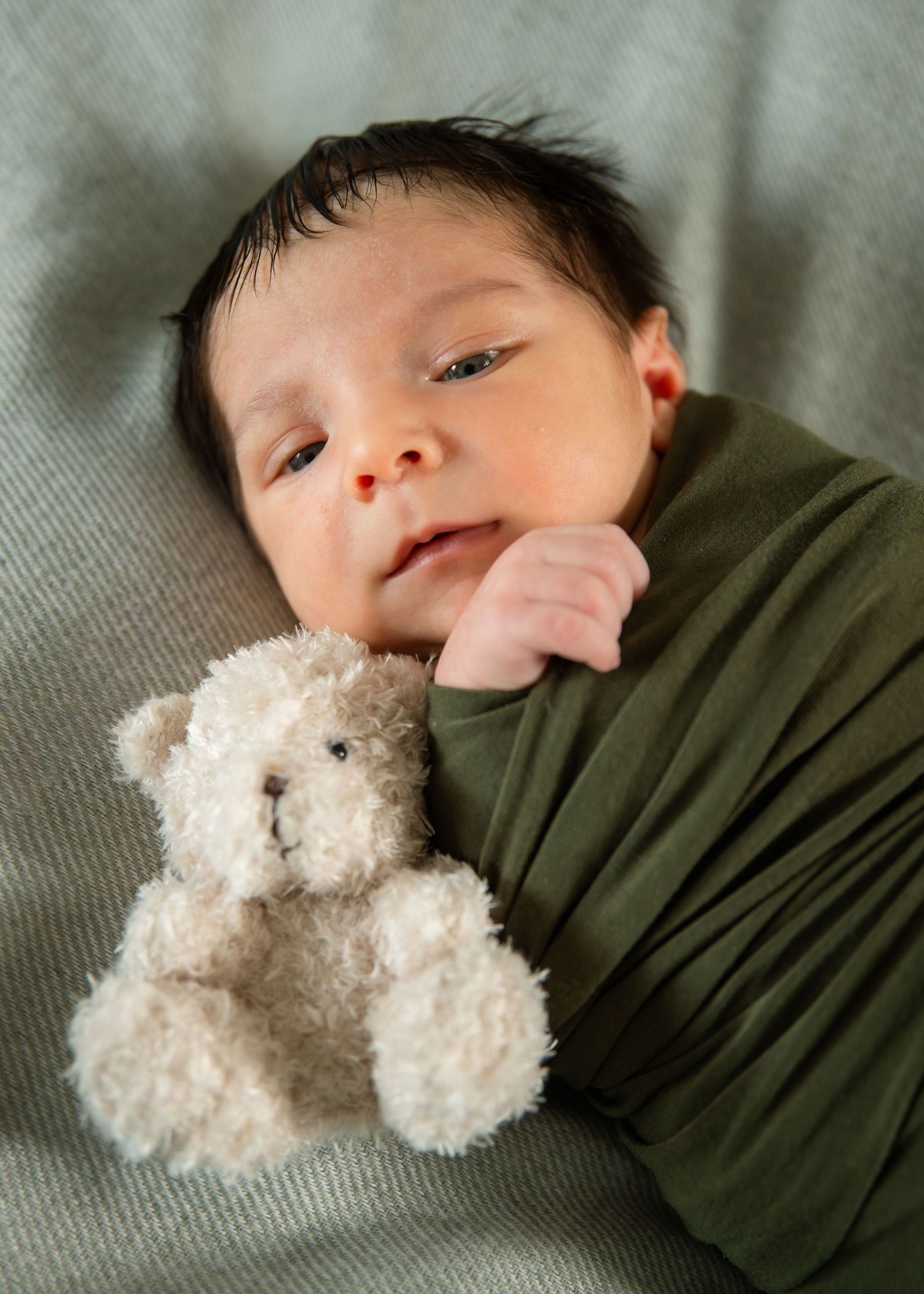 Newborn baby swaddled in green wrap lying on gray blanket with small teddy bear.