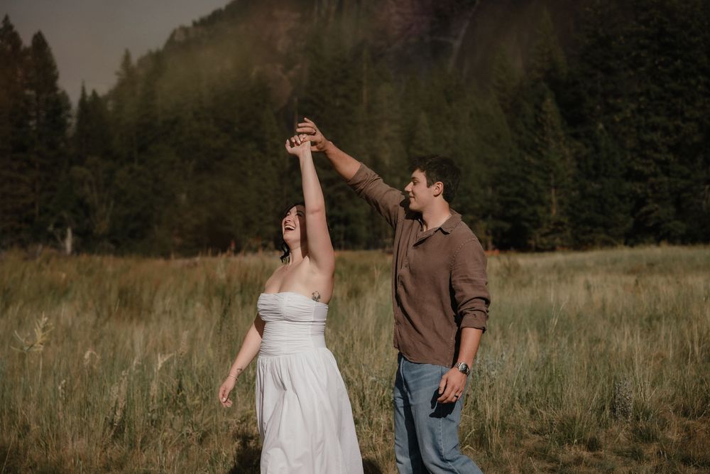 Couple dancing in a meadow with evergreen forest backdrop during golden hour engagement session.