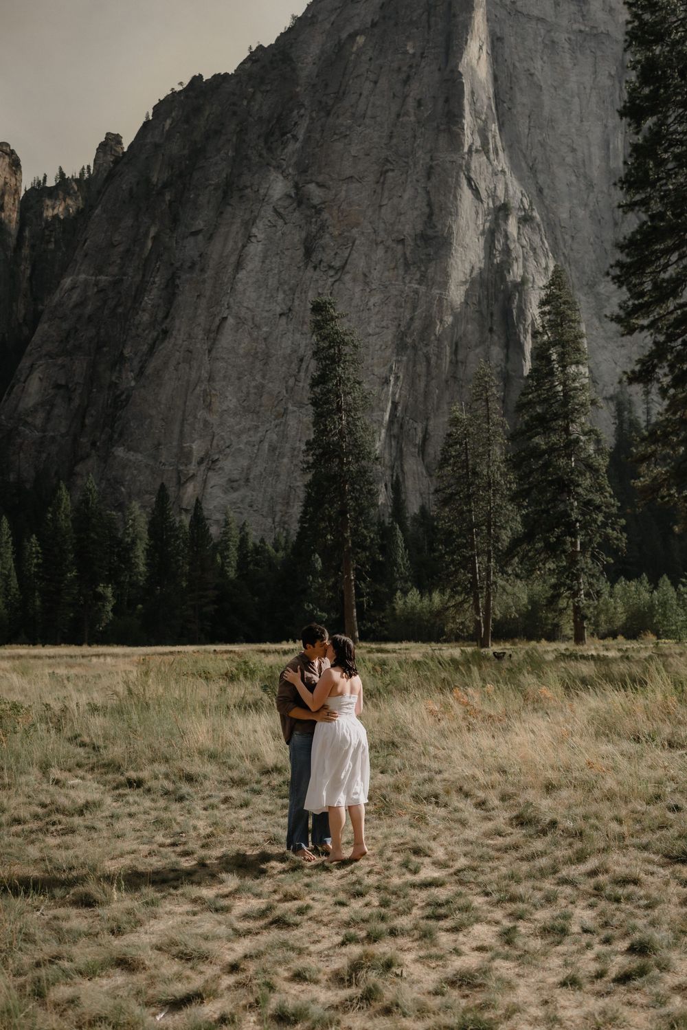 Couple embracing in Yosemite Valley meadow with towering granite cliff and pine trees backdrop.