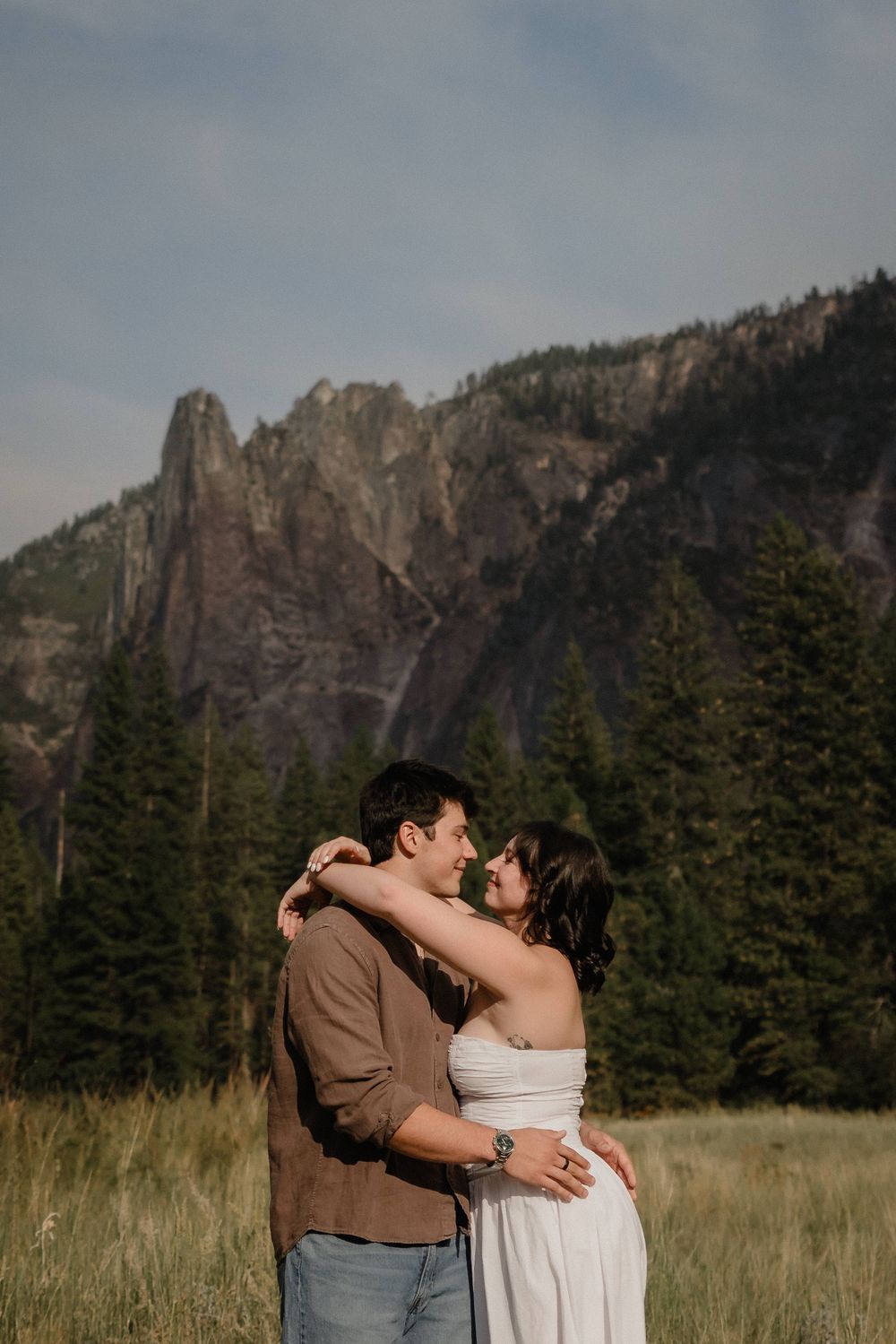Couple embracing in a meadow with dramatic mountain peaks and pine trees in Yosemite National Park.