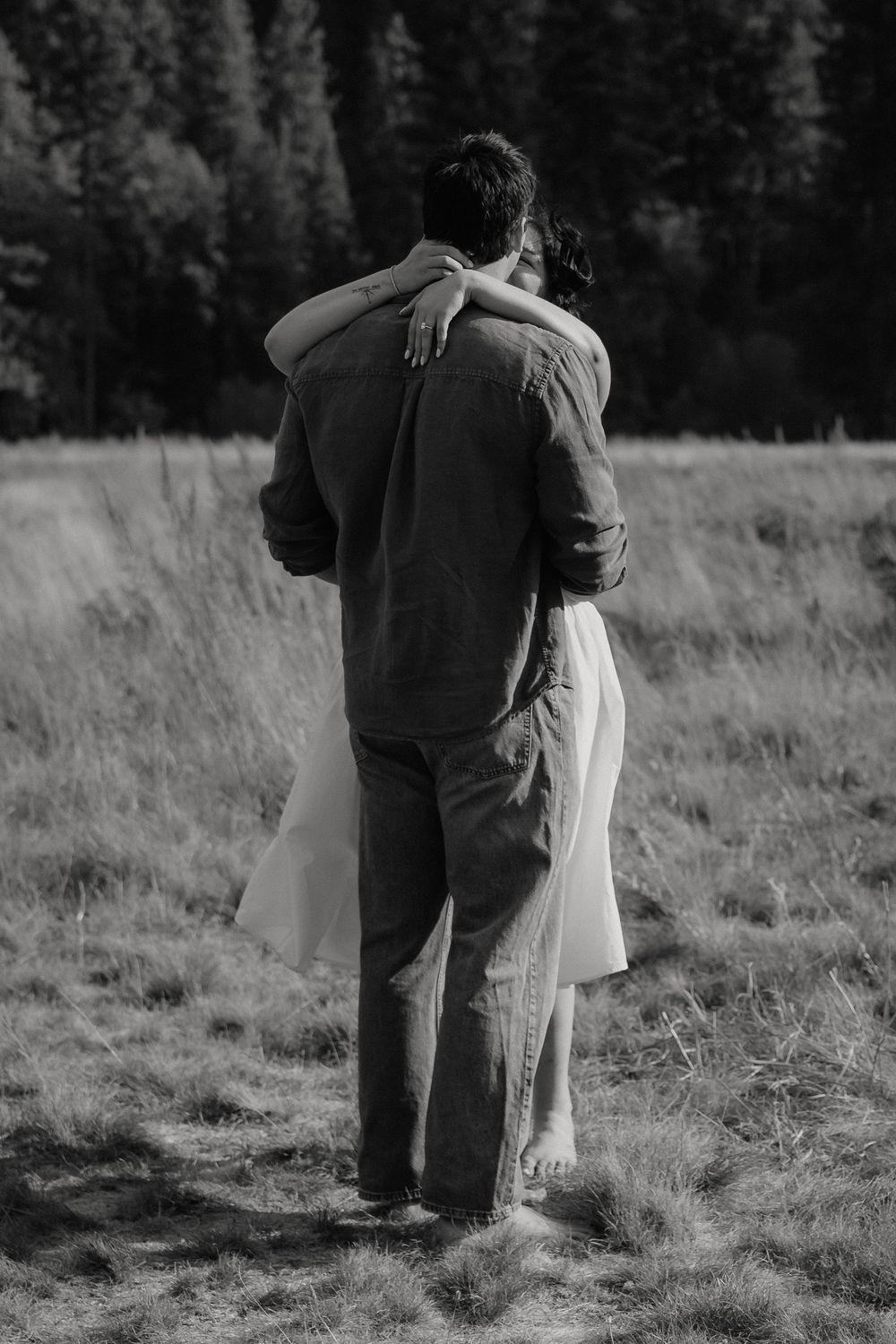 Black and white photo of a couple embracing in a meadow with forest backdrop, viewed from behind.