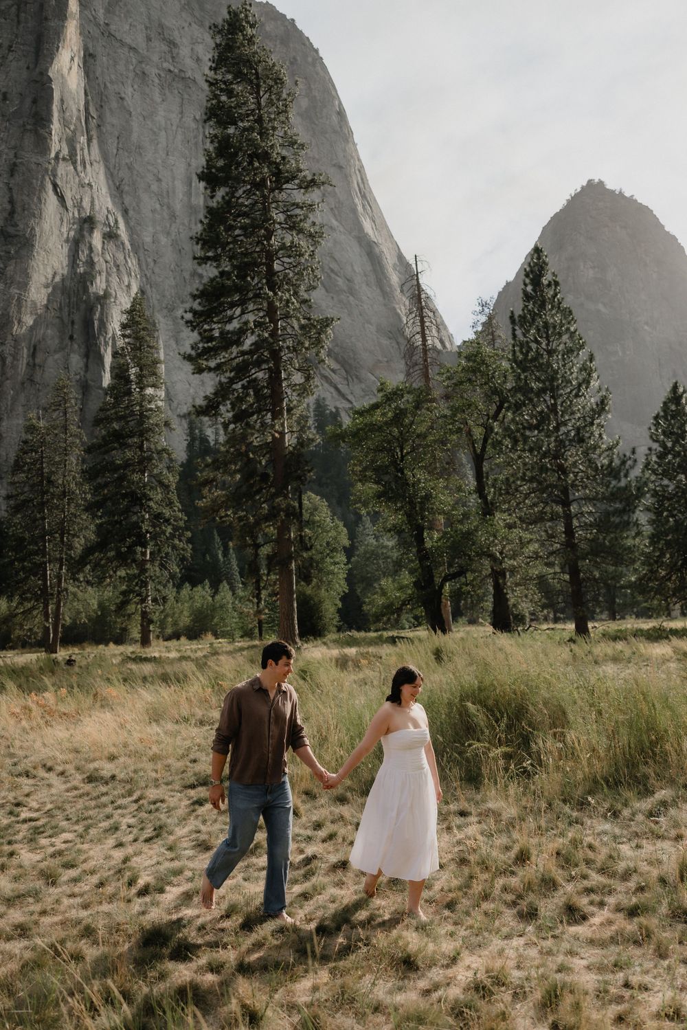 Couple holds hands walking through golden meadow with towering granite cliffs and pine trees in Yosemite National Park.