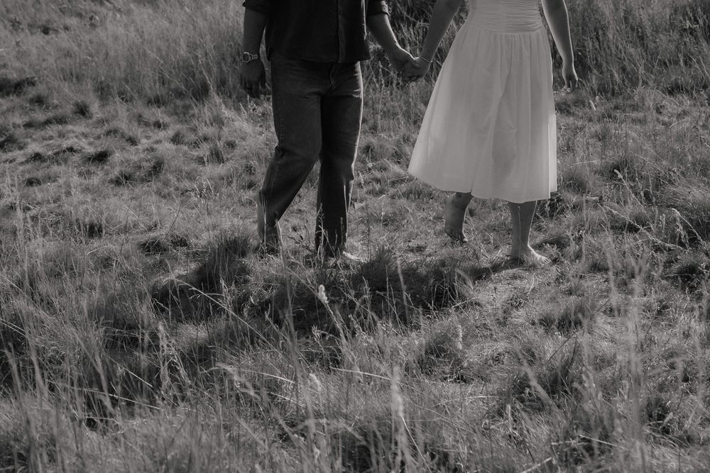Black and white photo of a couple holding hands walking barefoot through a grassy field.