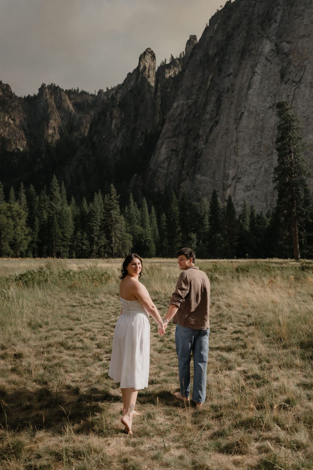 Couple holds hands in a meadow at Yosemite with dramatic granite cliffs and pine trees in the background.