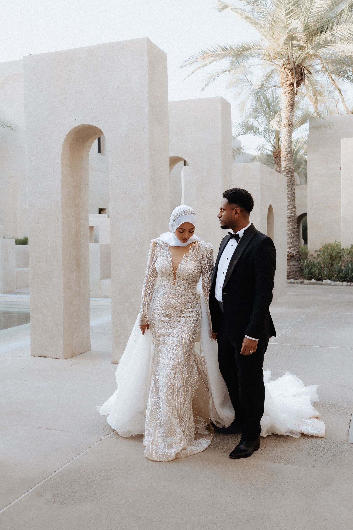 Bride in a hijab and groom in a tuxedo walking together among sandstone arches in Dubai