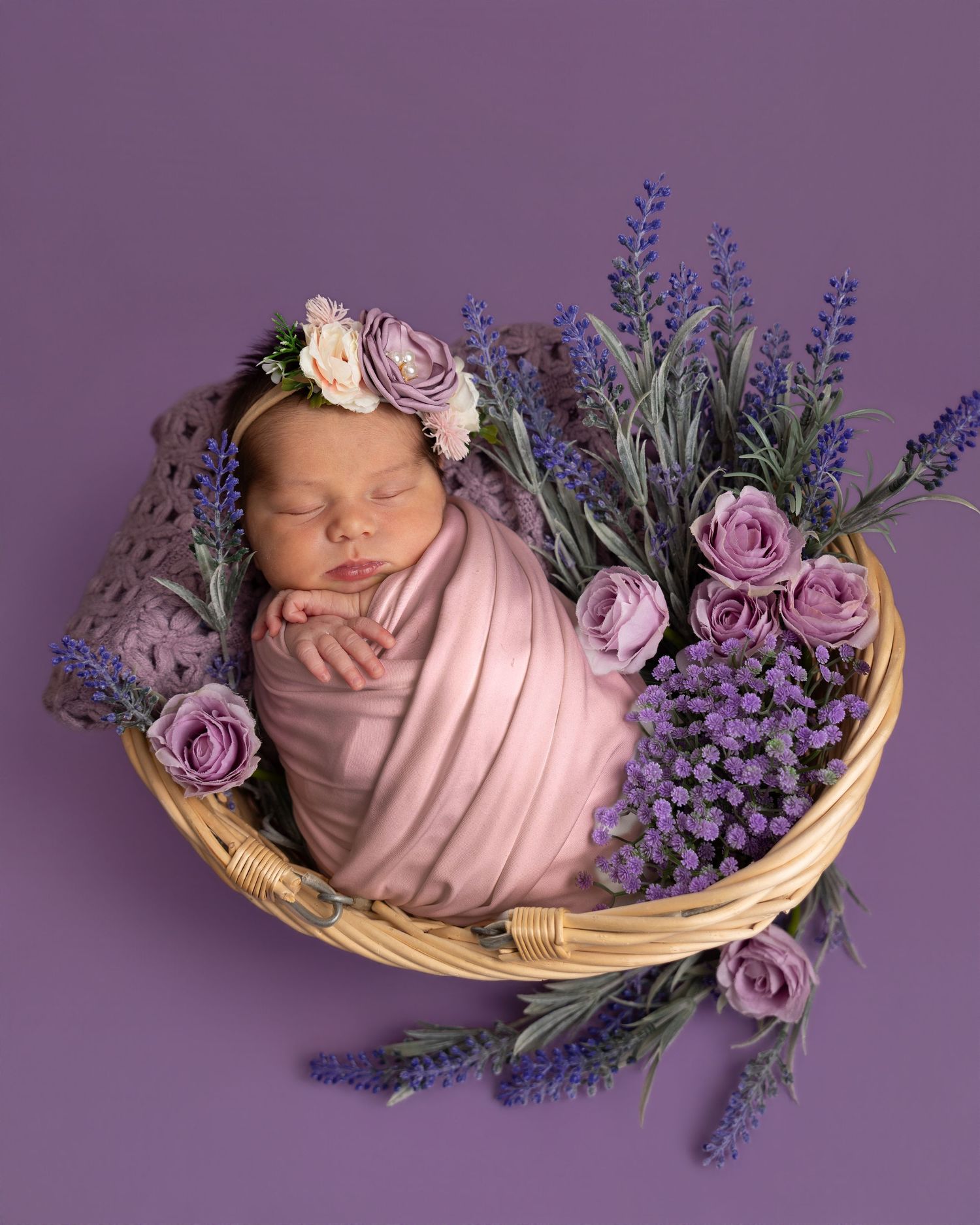 Newborn baby girl swaddled in pink wrap, sleeping in a wicker basket with purple lavender and roses.