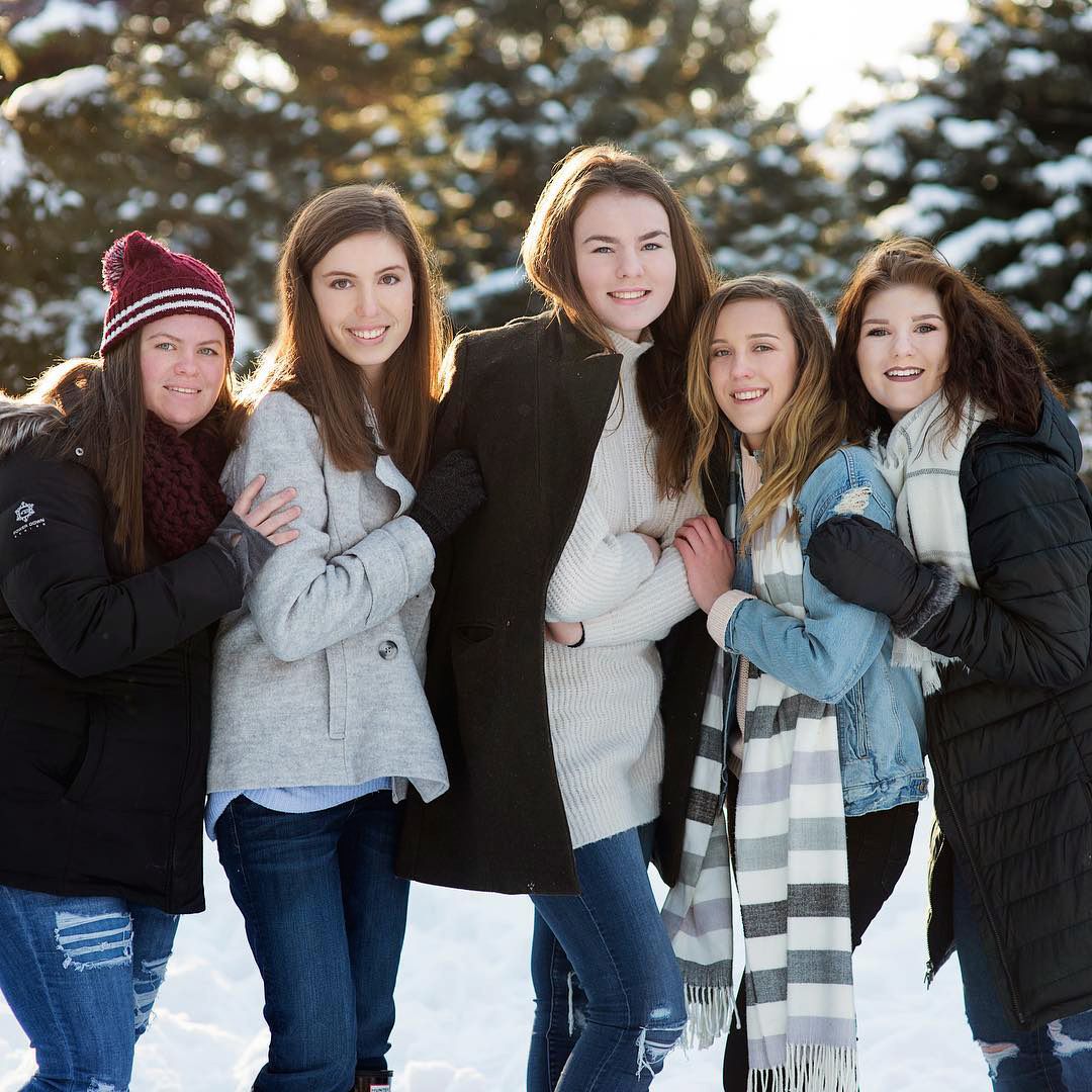 Five smiling teenage girls bundled in winter coats posing together in snowy woods.