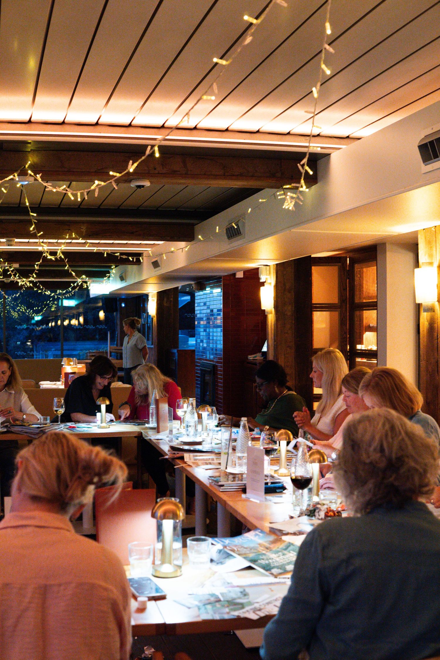 Guests dining at a warmly lit restaurant with fairy lights, long tables, and an inviting evening atmosphere.