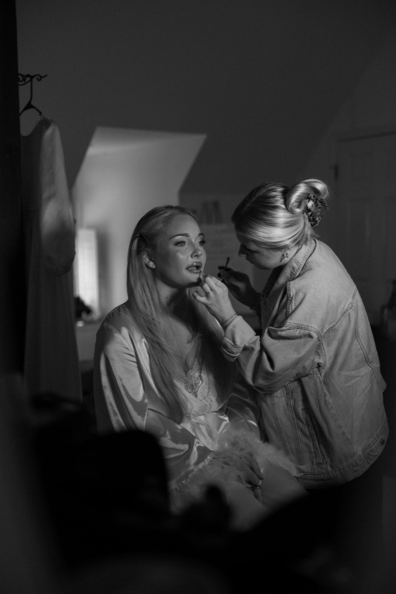 Black and white photo of a makeup artist applying lipstick to a bride in a satin robe during wedding preparations.