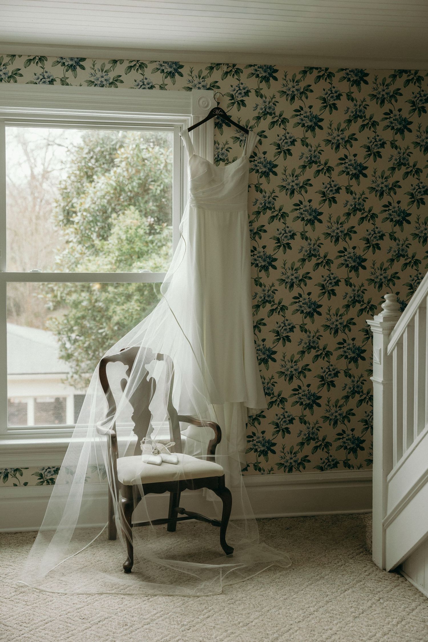 A white wedding dress with veil hangs near a window over a vintage chair in the getting ready space in a Georgia wedding venue.