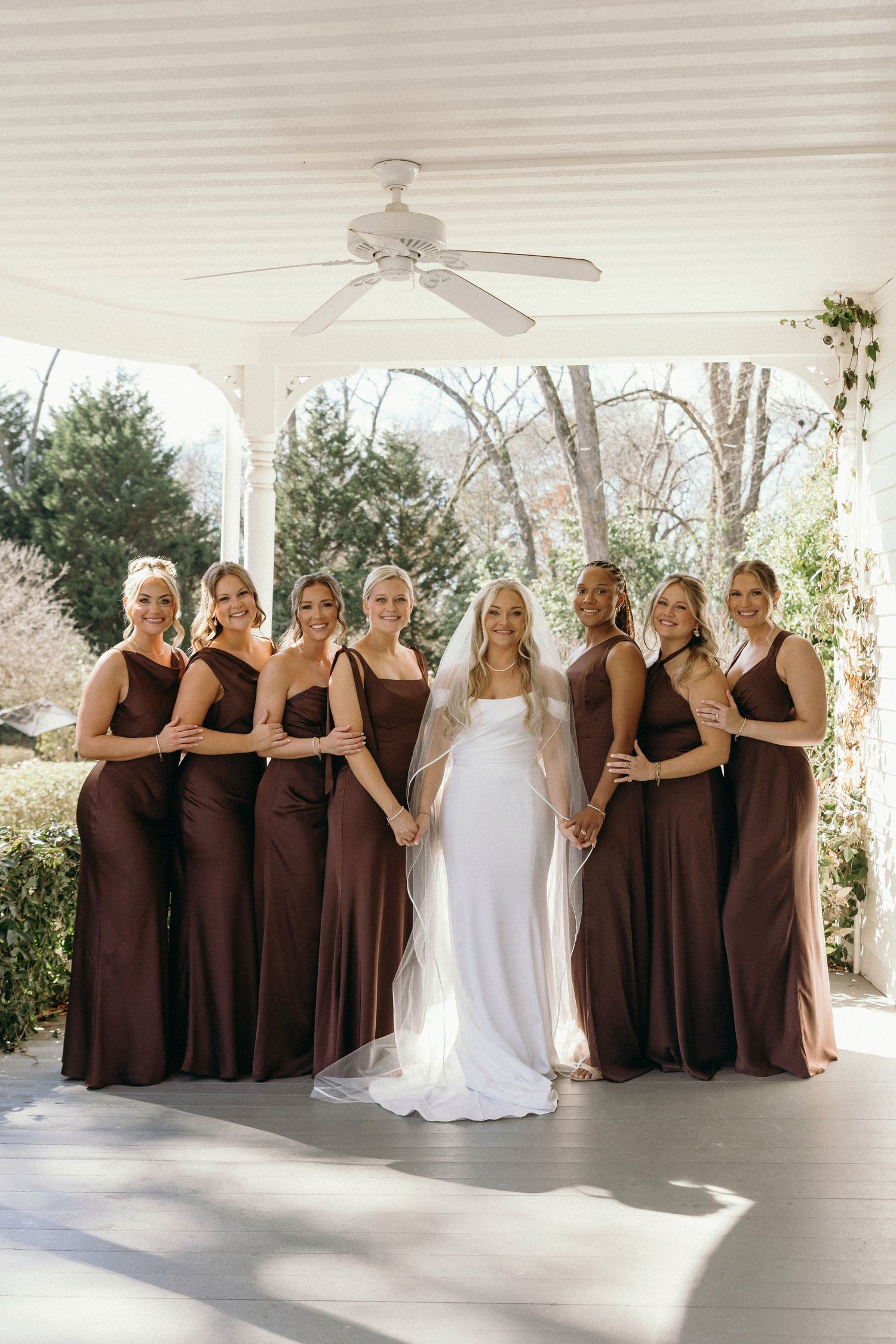 Bride in white gown with bridesmaids in brown dresses posing on porch at outdoor wedding venue.