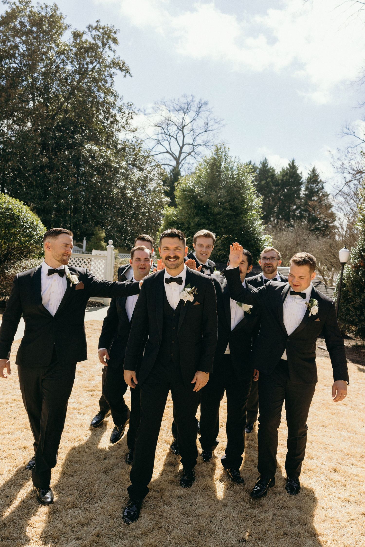 Groom and groomsmen in black tuxedos with bow ties walk outdoors at a wedding venue surrounded by trees.