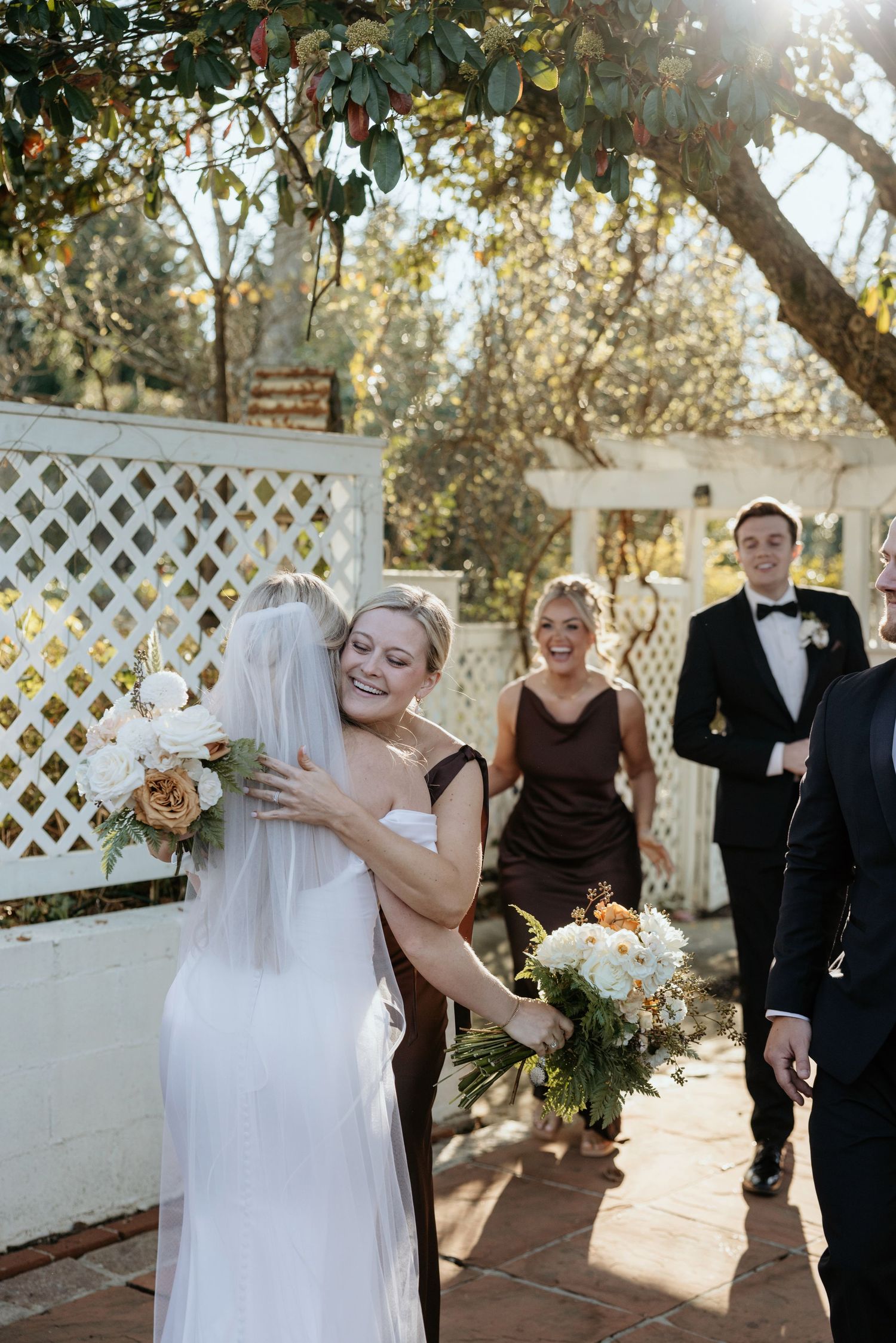Bride and guest embrace joyfully outdoors at a wedding, with bridesmaids and groomsmen smiling in the background.