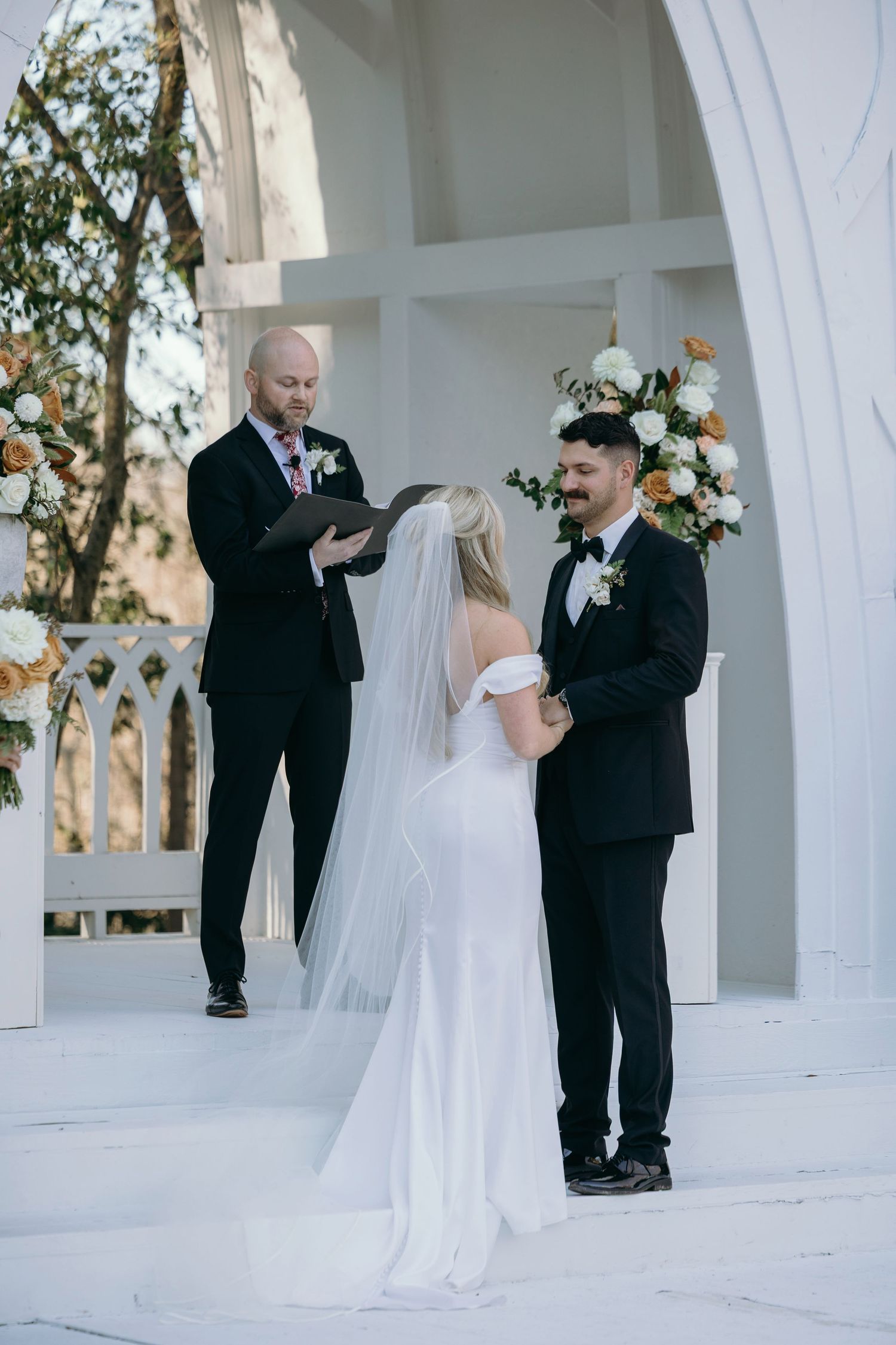 Bride and groom exchanging vows at an elegant white outdoor wedding ceremony with floral arrangements.