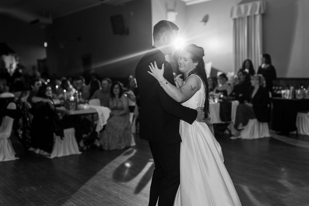Bride in a white gown and groom in a dark suit share a first dance in a ballroom while wedding guests watch from tables.