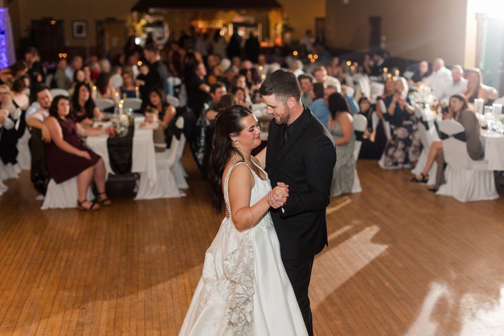 Bride in a white gown and groom in a black suit share a first dance on a wood floor while wedding guests watch from nearby tables.