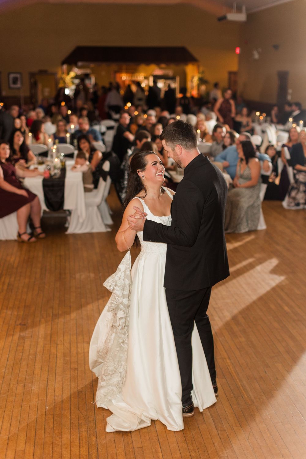 Smiling bride in a white gown and groom in a black suit share a first dance on a wood floor while seated wedding guests watch.