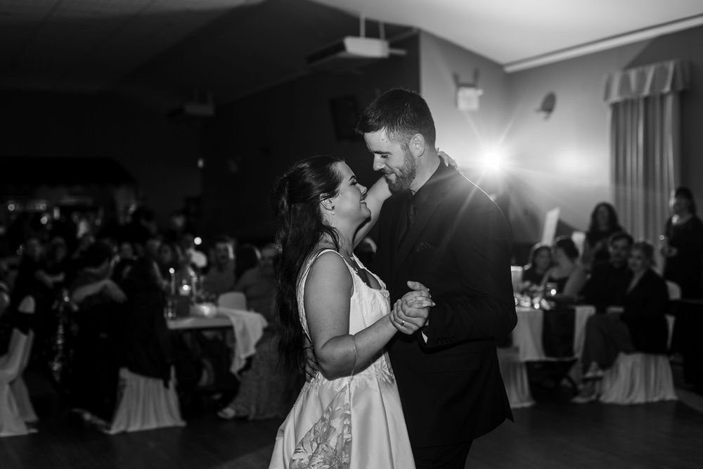 Bride in a white gown and groom in a dark suit share a slow dance while wedding guests watch from tables in the background.