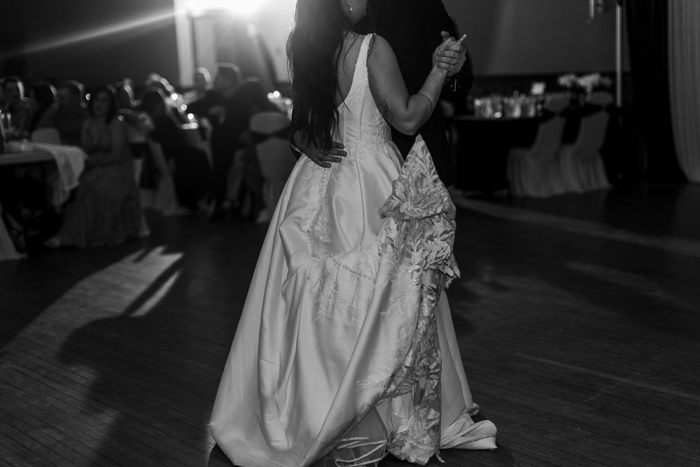 Black and white photo of a bride in a flowing satin gown with lace detail dancing at a wedding reception as guests watch.